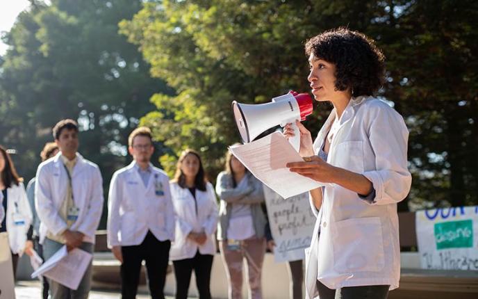 Sheyda Aboii speaks to her fellow medical students, including, background left to right, Arun Burra, Andrés Calvillo and Avery Thompson and the community, at a White Coats for Black Lives (WC4BL) Teach-in, which set out to “model an explicitly anti-racist healthcare education that elevates queer, disabled, formerly criminalized, and undocumented people of color in the communities and hospitals within which we work,” on the 4th anniversary of WC4BL in 2018. Photo by Susan Merrell.